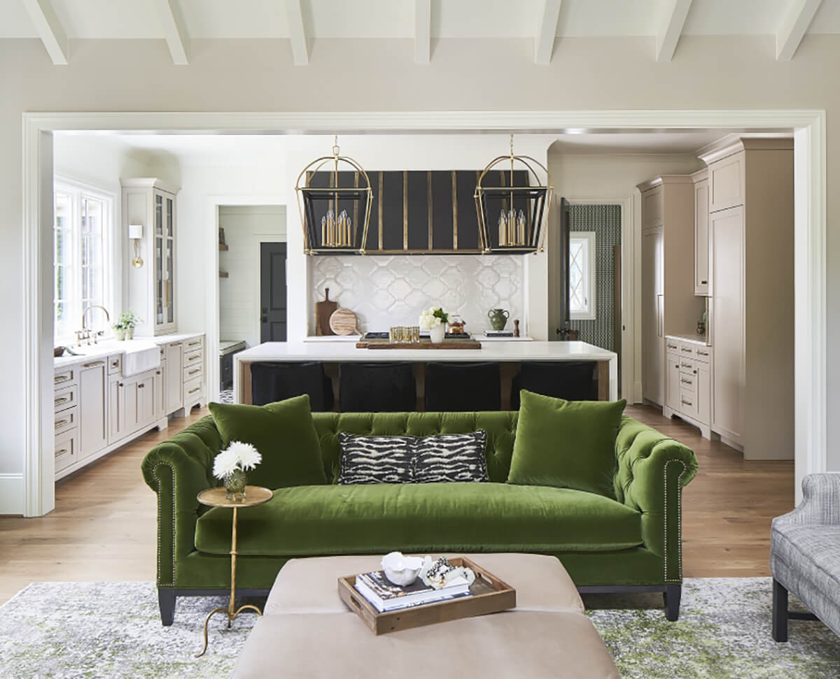 A Southern moern farmhouse kitchen with soft, off-white cabinets, a black metal hood, dark gray kitchen island, and olive green accents including the green couch in the neighboring living room.